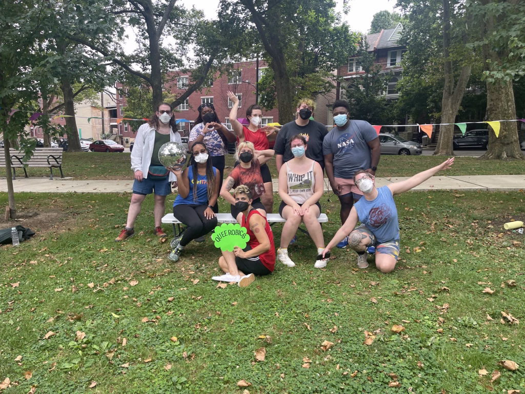 A masked group of Queerobics participants outdoors, lined up behind a large, multicolored 