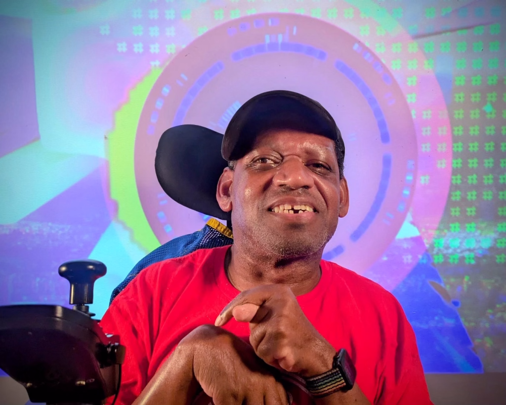 Man in power wheelchair, wearing a black hat and a red shirt, smiles at the camera with a colorful background.