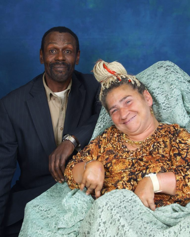 This is a picture of Pamela Price and her friend Eric. Pam is a white person wearing an orange and black shirt and gold necklace and earrings. Her hair is adorned with white braids pulled up into a bun on top of her head — one braid in the front is red. Pam is also wearing a turquoise blanket. Eric, a Black man, is wearing a tan shirt and dark blue suit jacket. Both Eric and Pam are smiling wide.