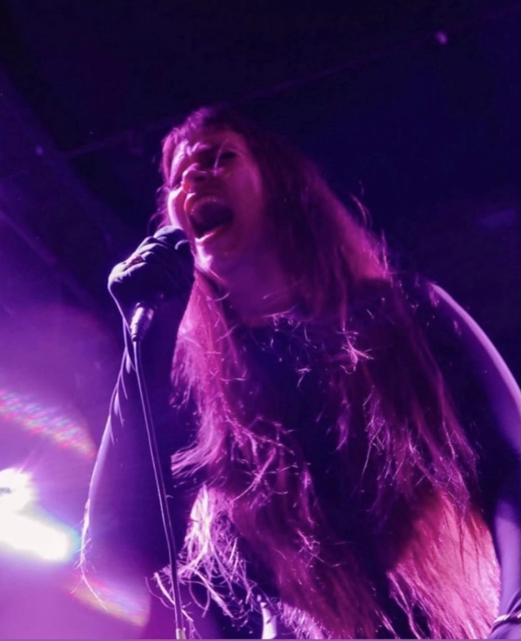 a picture of Anna singing into a microphone wearing an all black gloved body suit. Bathed in purple lighting. Photo by Rachael Warriner.