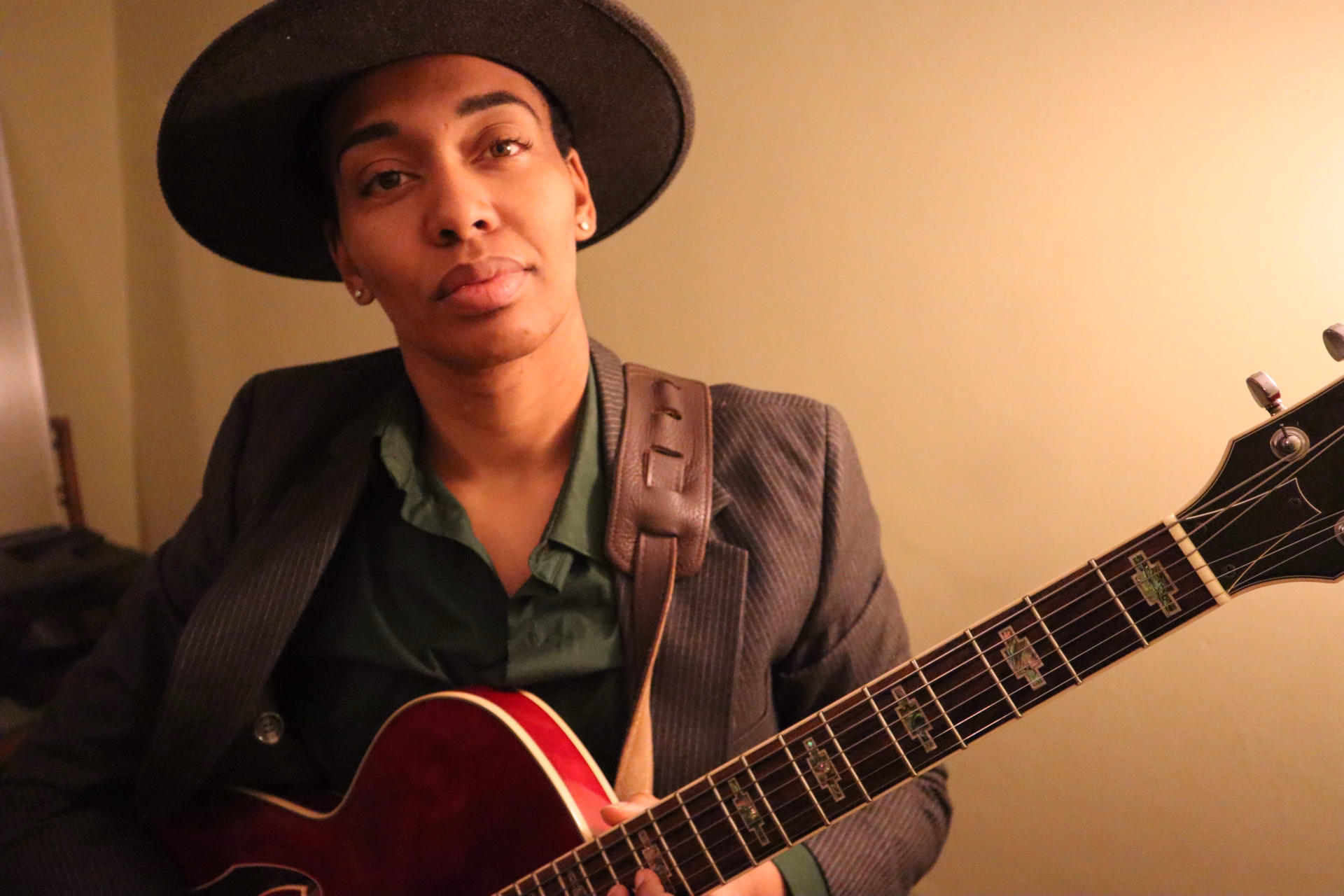 Ash Phillips is pictured holding a red, semi-hollow bodied guitar with a leather guitar strap. They are wearing a grey, wide-brimmed hat, studed earrings in each ear, a green button-up up shirt, and pin-striped blazer. They are looking intently towards the camera.
