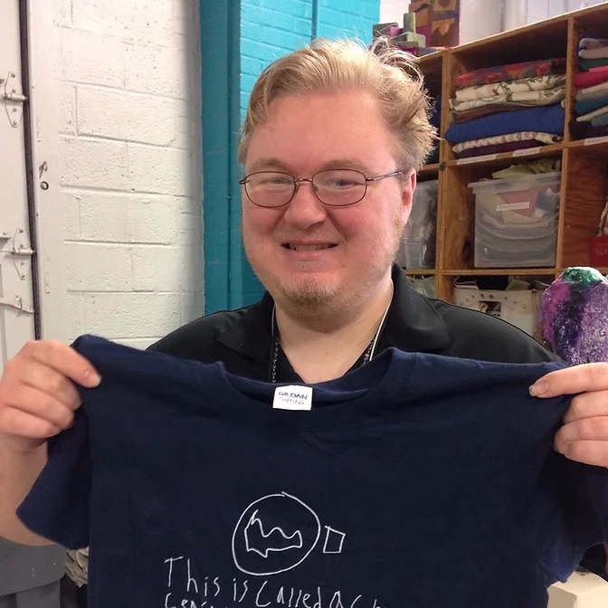 Owen smiles at the camera and holds up a tshirt with his design on it.
